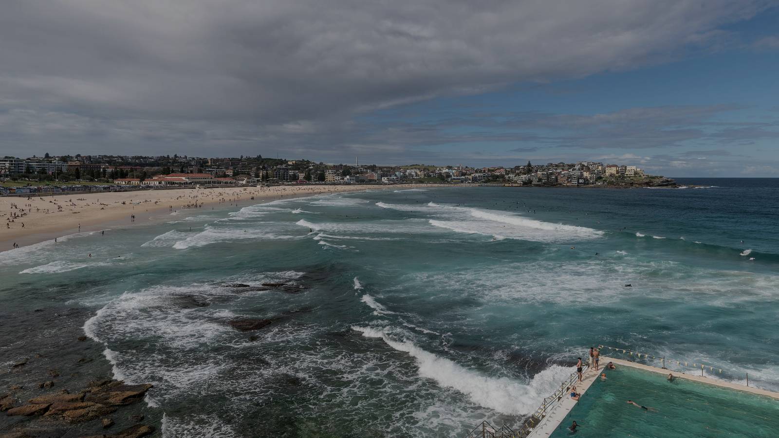 Waves rolling onto Bondi Beach, Sydney, under a cloudy sky, with the Bondi Icebergs ocean pool in the foreground.
