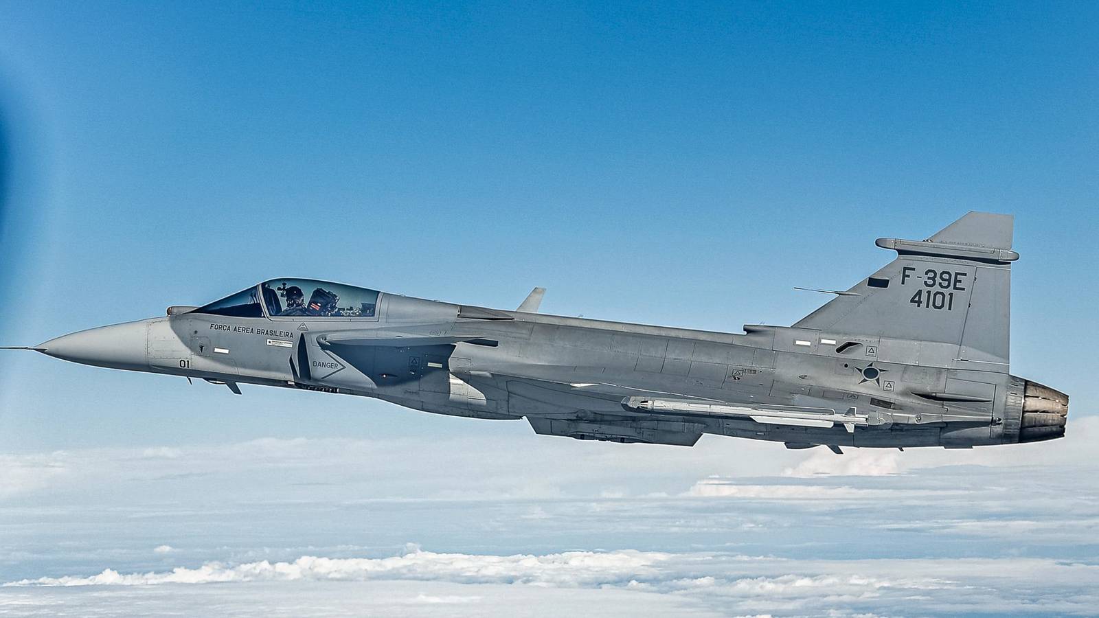 A grey Brazilian Air Force F-39E Gripen fighter, tail number 4101, flying above a layer of white cloud under a blue sky