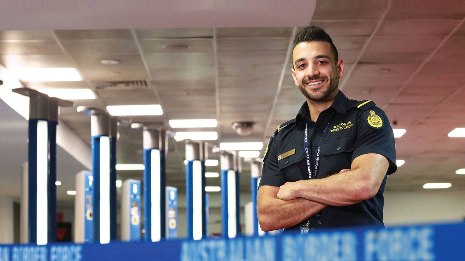 An Australian Border Force officer in uniform smiling with arms folded in an airport arrivals area