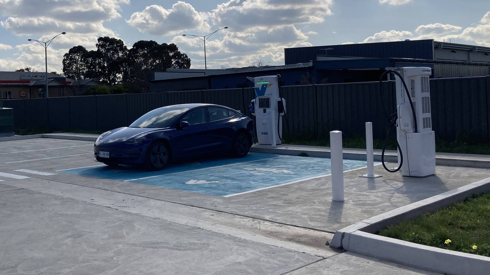 A blue electric car parked in a marked bay beside two Evie Networks fast chargers in an open car park