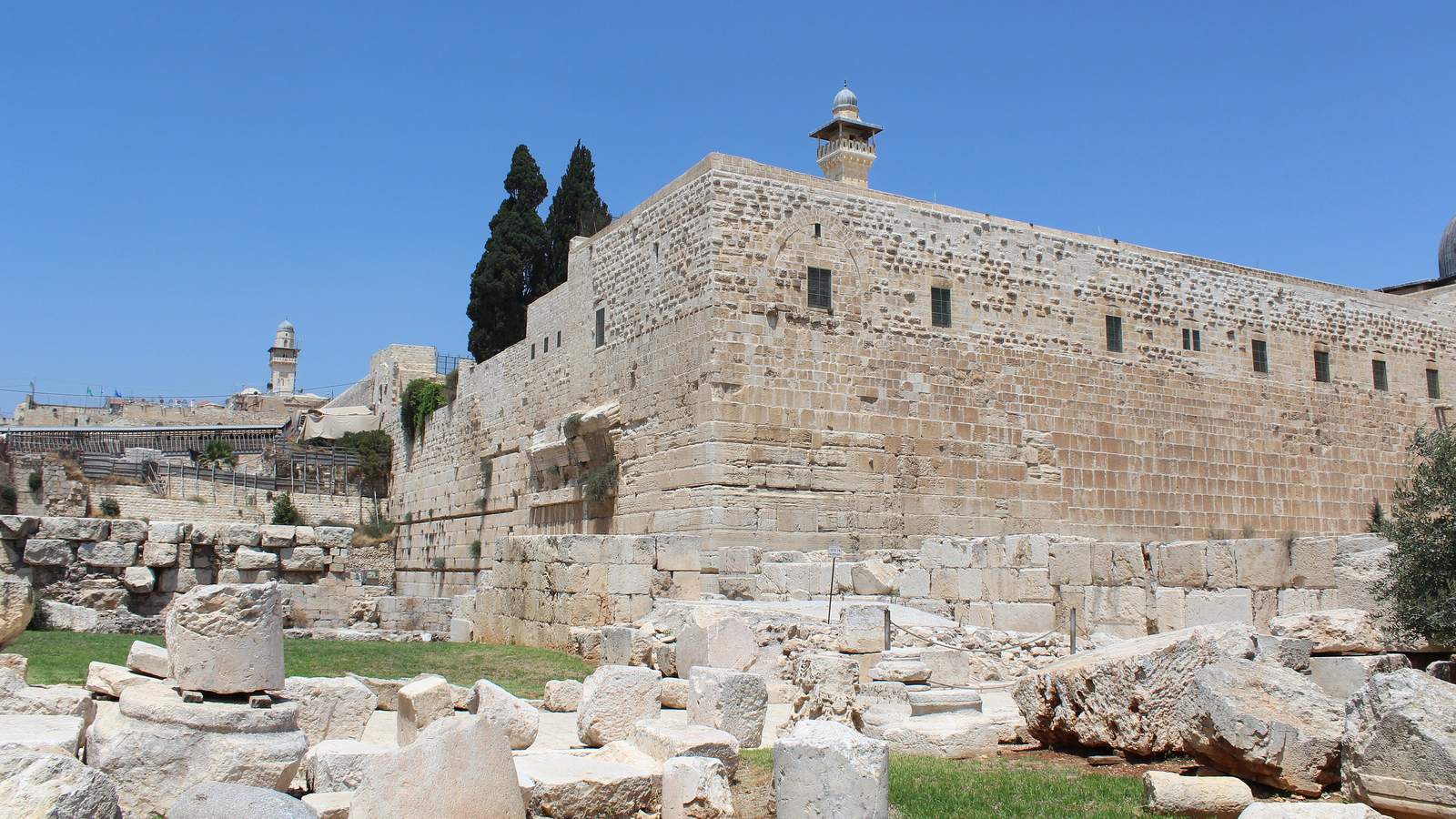 The Southern Wall of Jerusalem's Old City under a blue sky, with ancient fallen stones and cypress trees in the foreground
