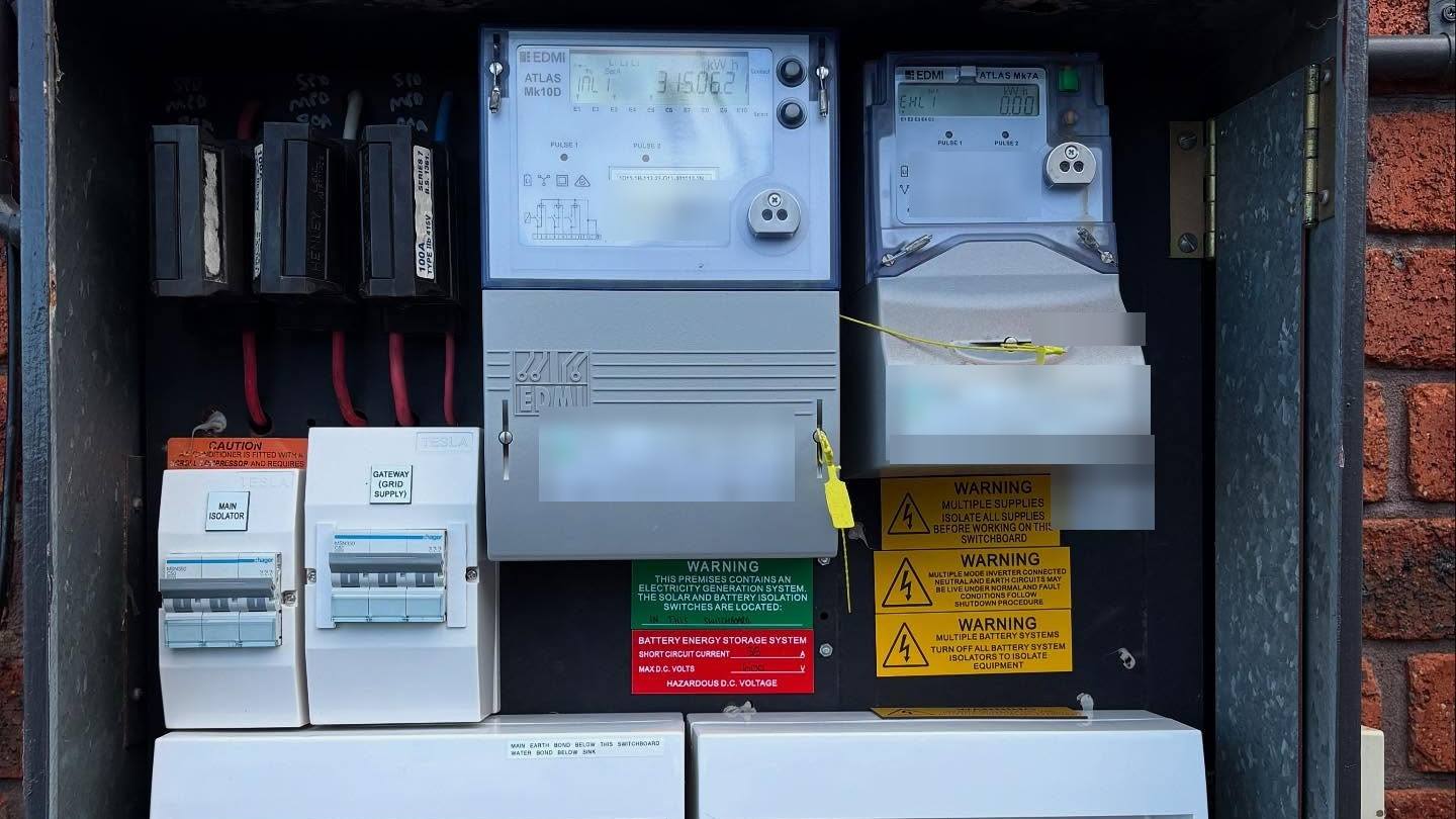 An open meter box on a brick wall with two electricity meters, isolator switches and yellow battery system warning labels