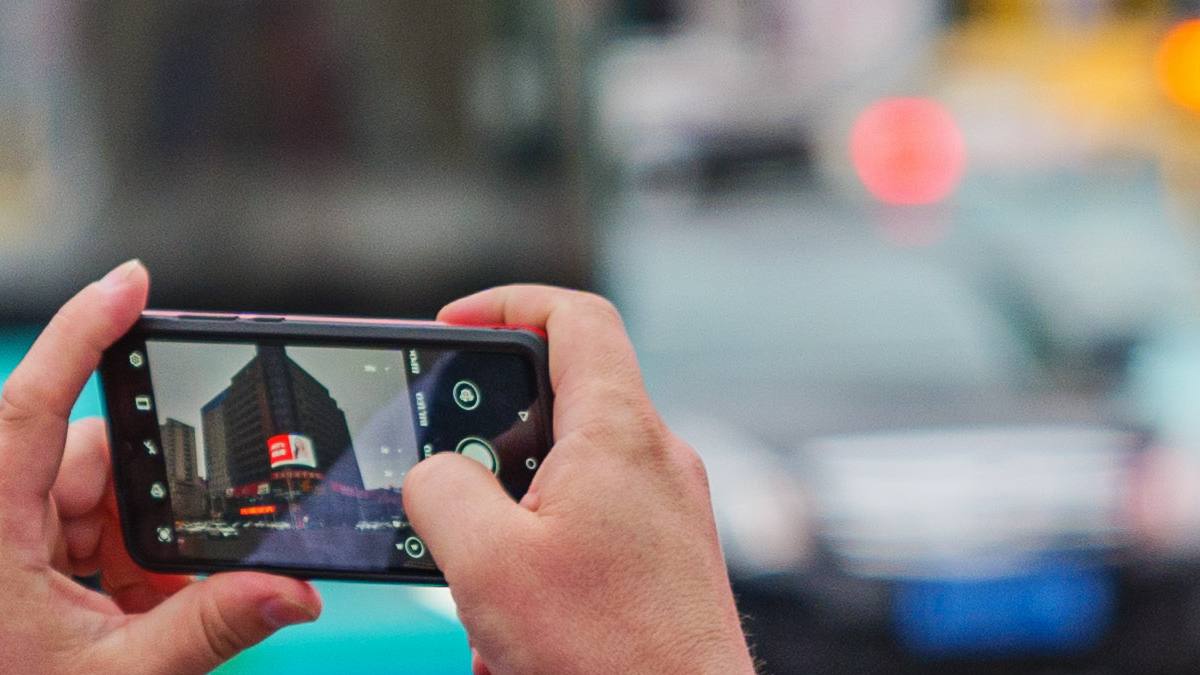 Two hands holding a smartphone sideways to take a photo of a city street, the camera app showing buildings on screen