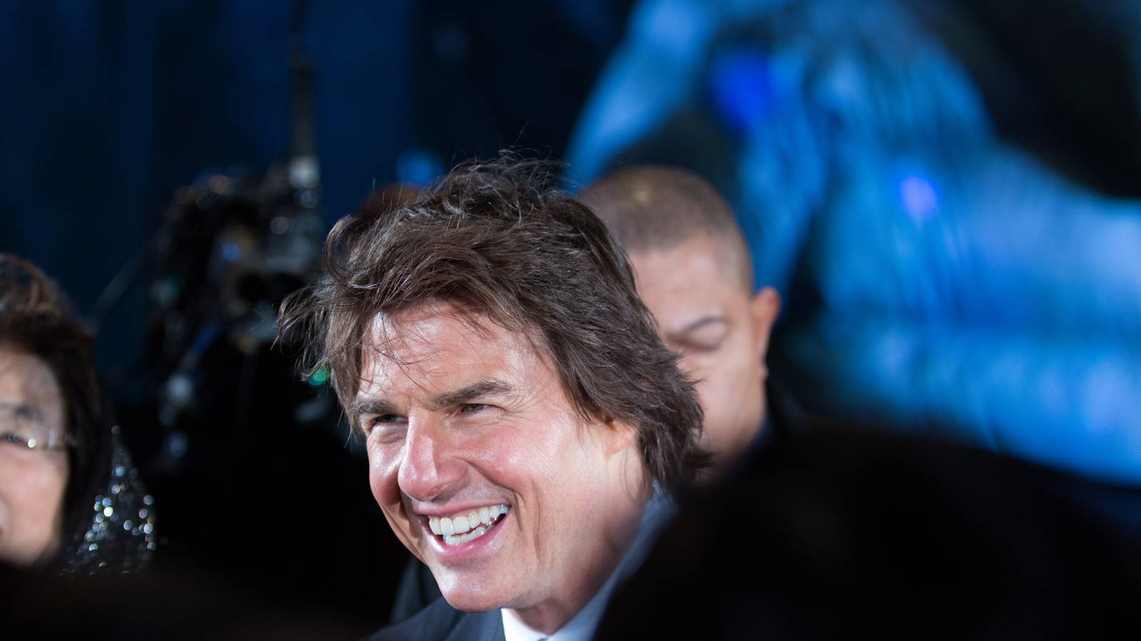 Tom Cruise waves to fans on a red carpet at a film premiere, wearing a dark suit