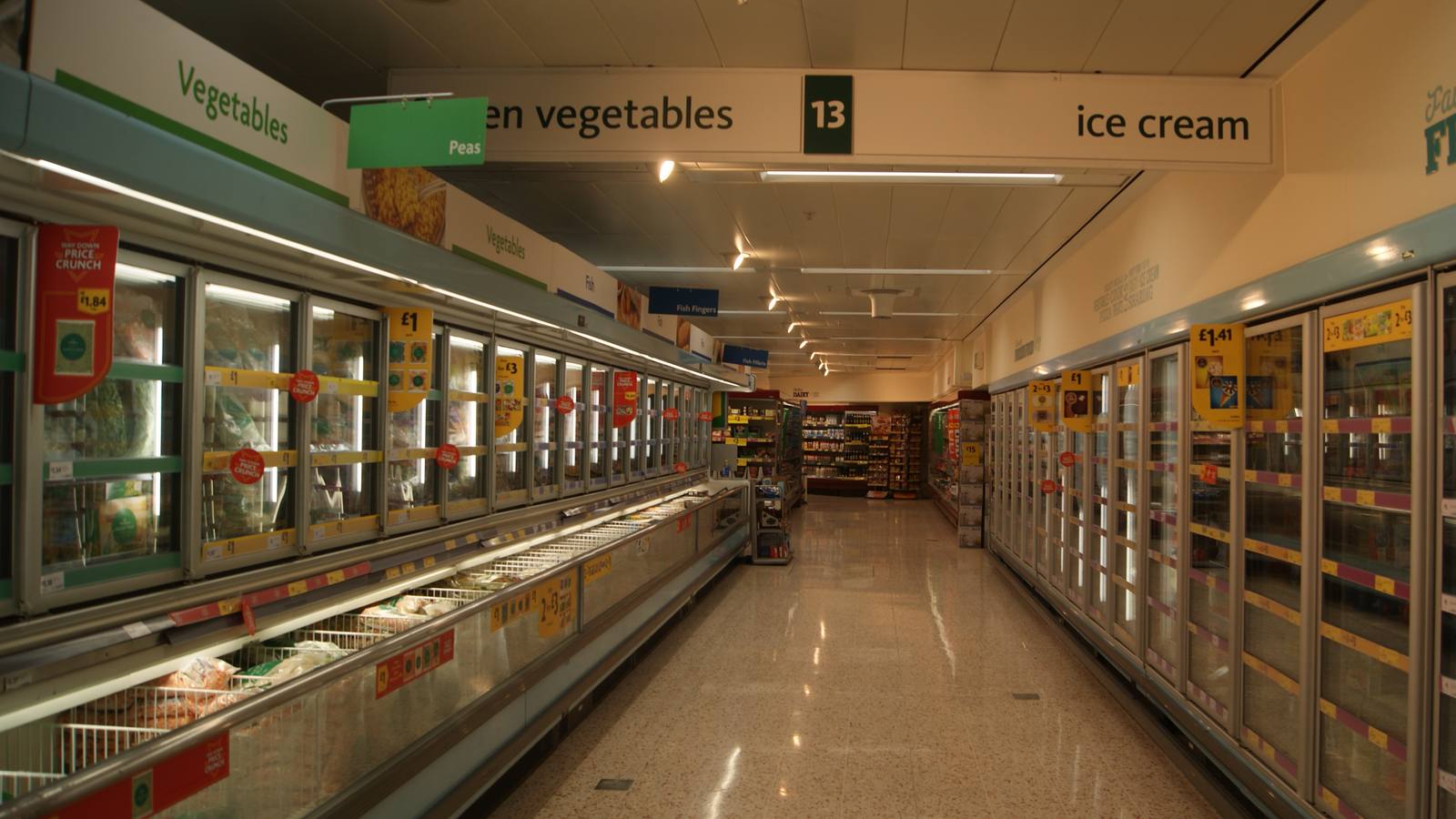 A long supermarket frozen food aisle lined with glass-door freezers under signs for vegetables and ice cream