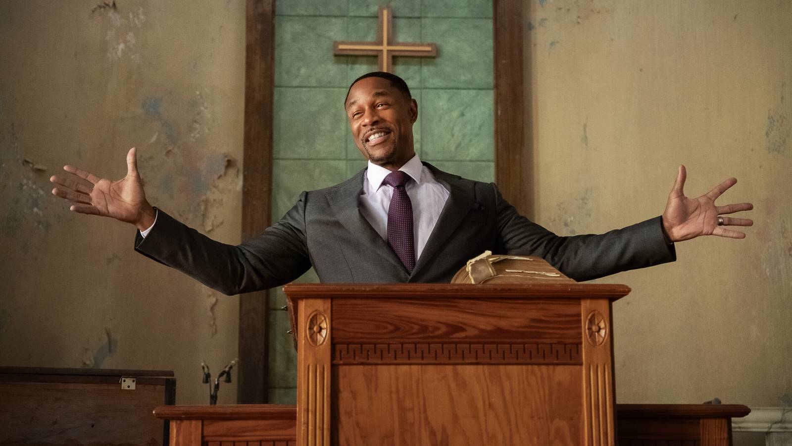 Tank as Pastor Isaac spreads his arms wide behind a wooden pulpit in a church