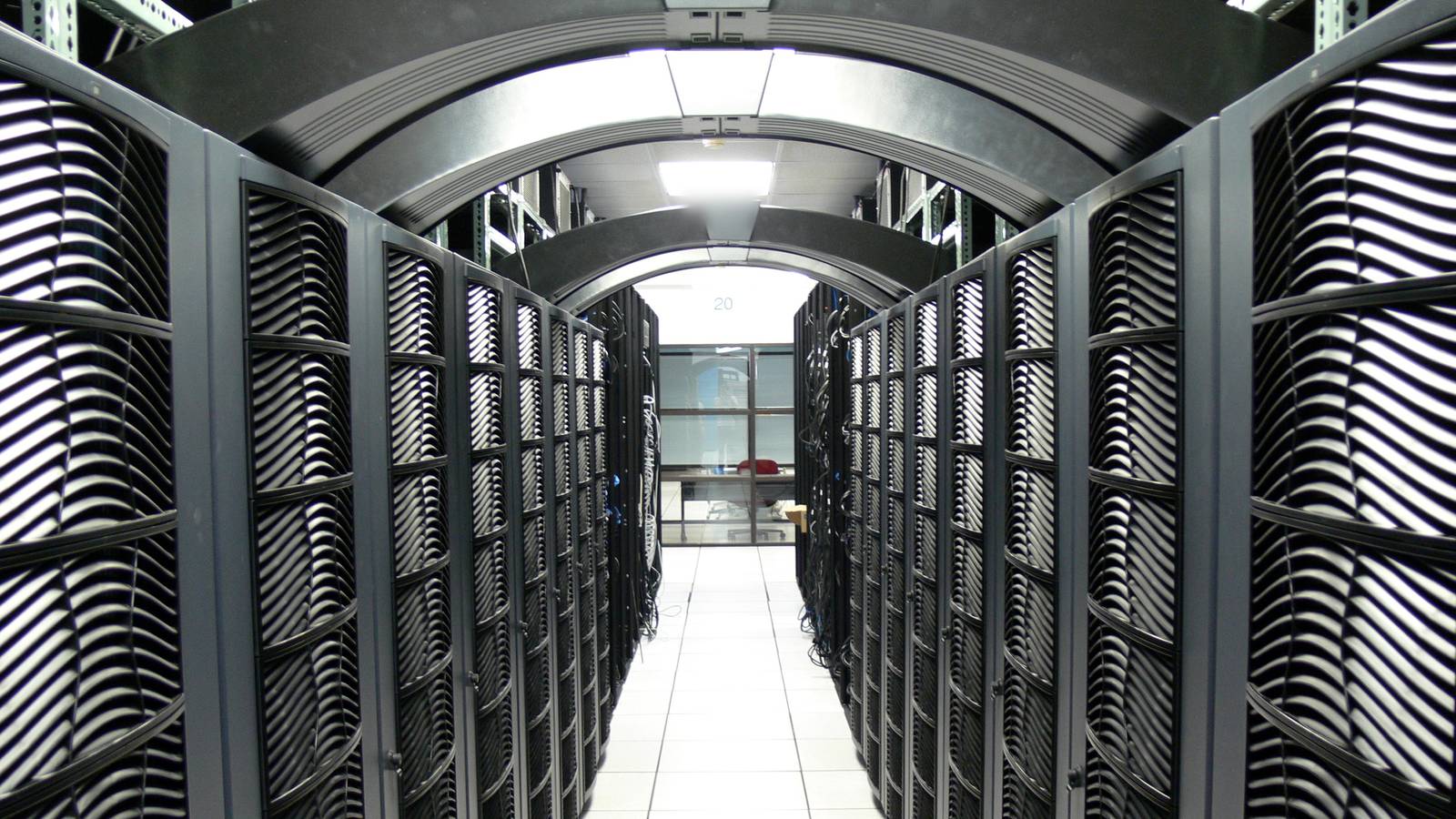 A long aisle between rows of black server cabinets in a brightly lit data centre