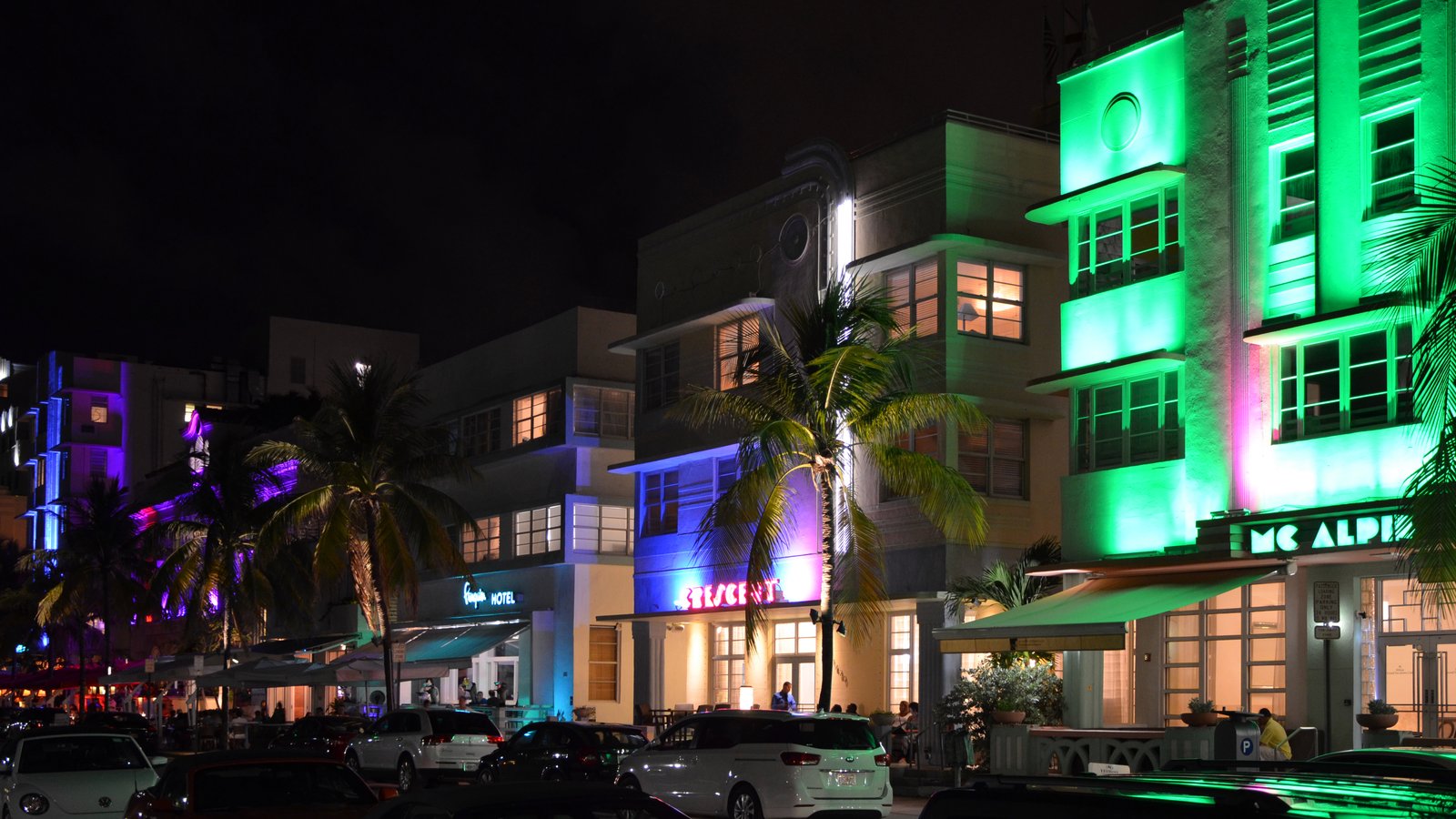 Art Deco hotels on Ocean Drive, Miami Beach, lit up in green, purple and blue neon at night, with palm trees and parked cars
