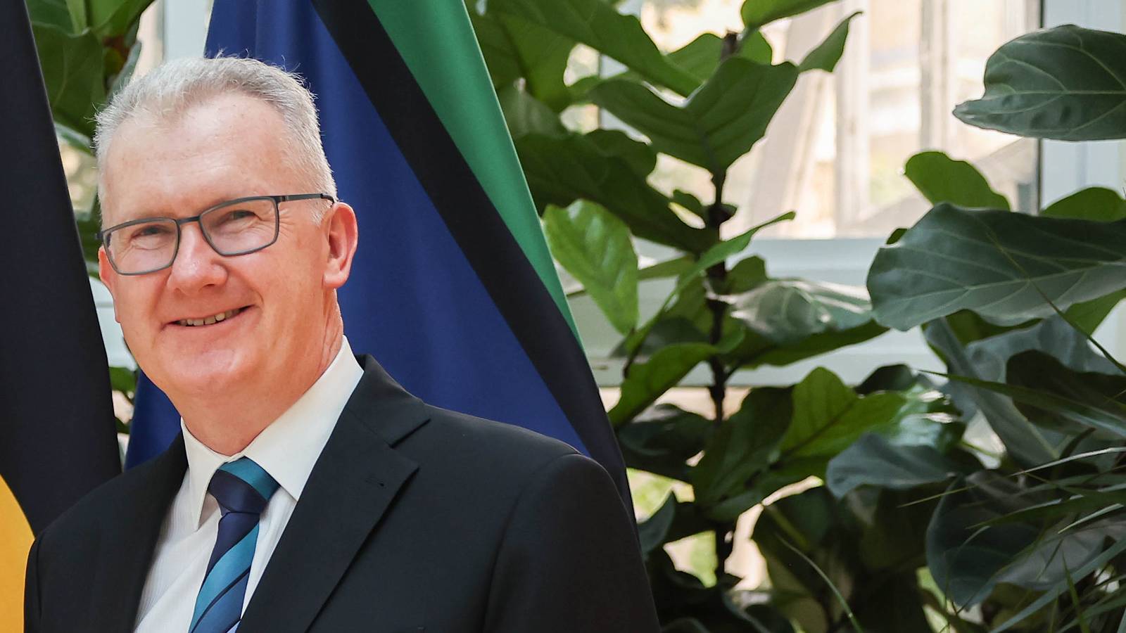 Home Affairs Minister Tony Burke, smiling in a dark suit and blue striped tie, in front of flags and indoor plants