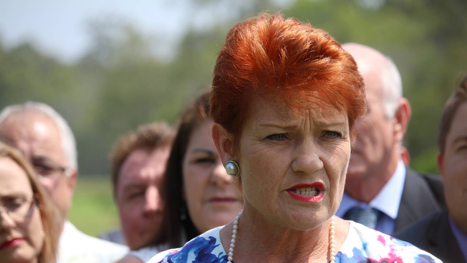 Pauline Hanson, with red hair and a floral dress, speaks outdoors with supporters standing behind her