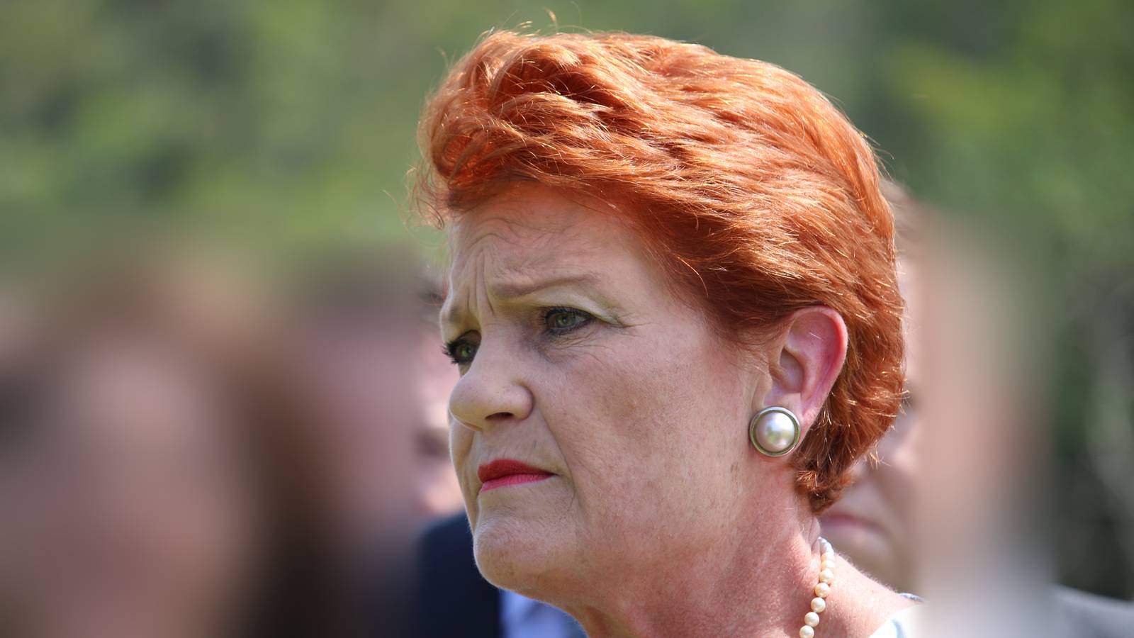 Pauline Hanson with short red hair, pearl earrings and a pearl necklace, looking to one side with a serious expression