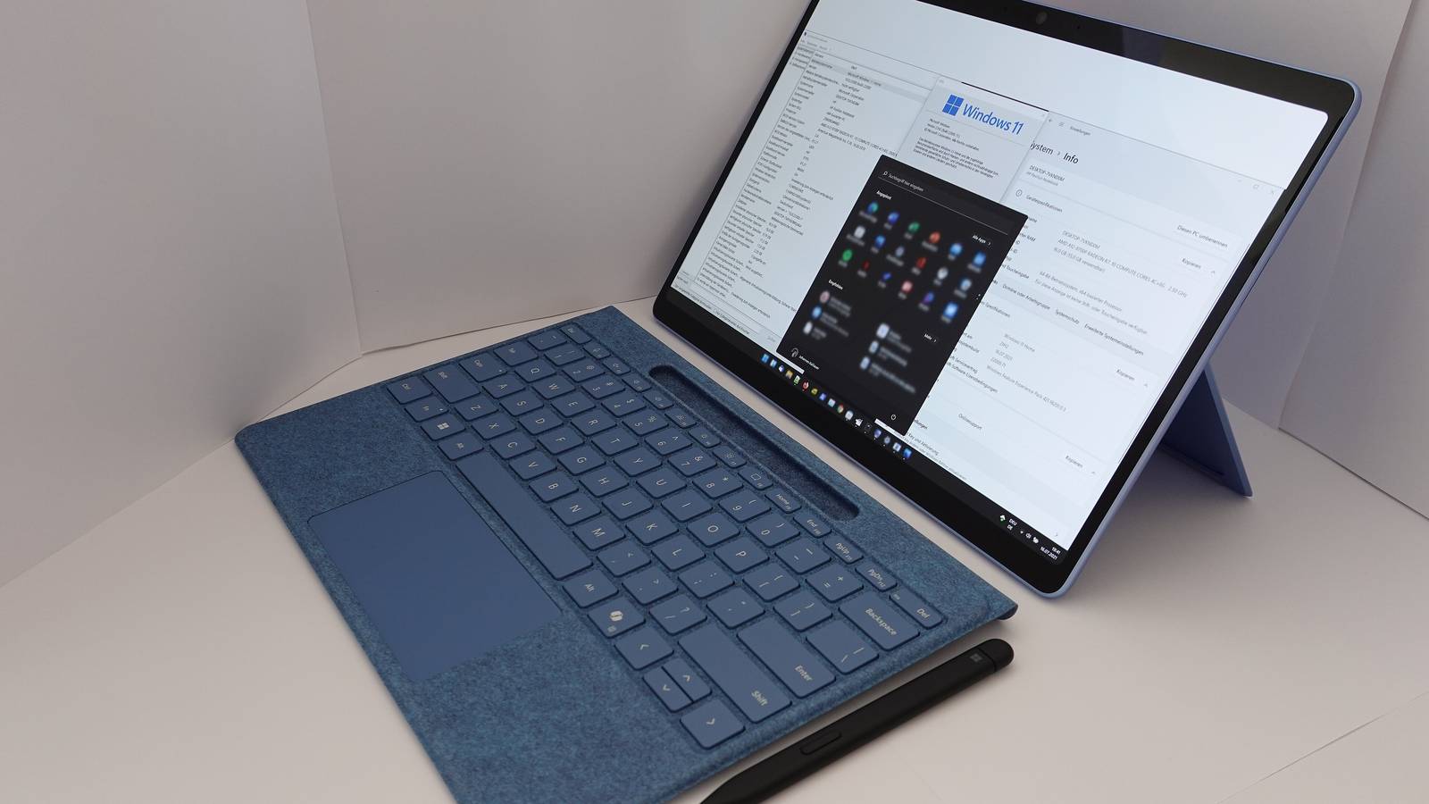 A Surface tablet with a blue keyboard cover and pen showing Windows 11 settings and the Start menu