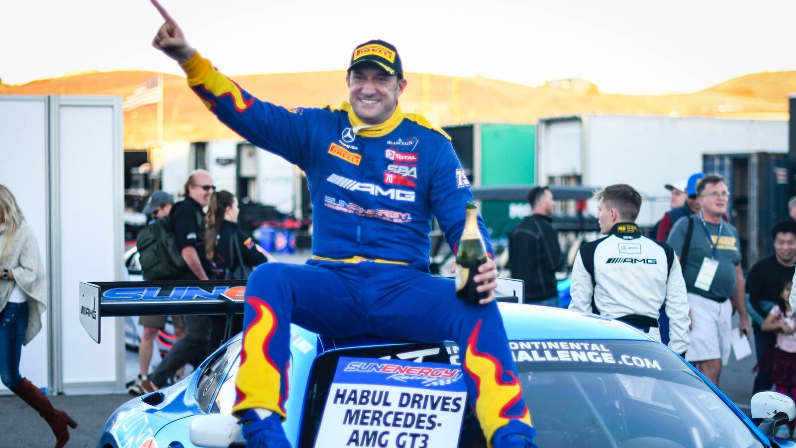 Kenny Habul in blue race overalls sits on his Mercedes AMG GT3 holding champagne and pointing skyward