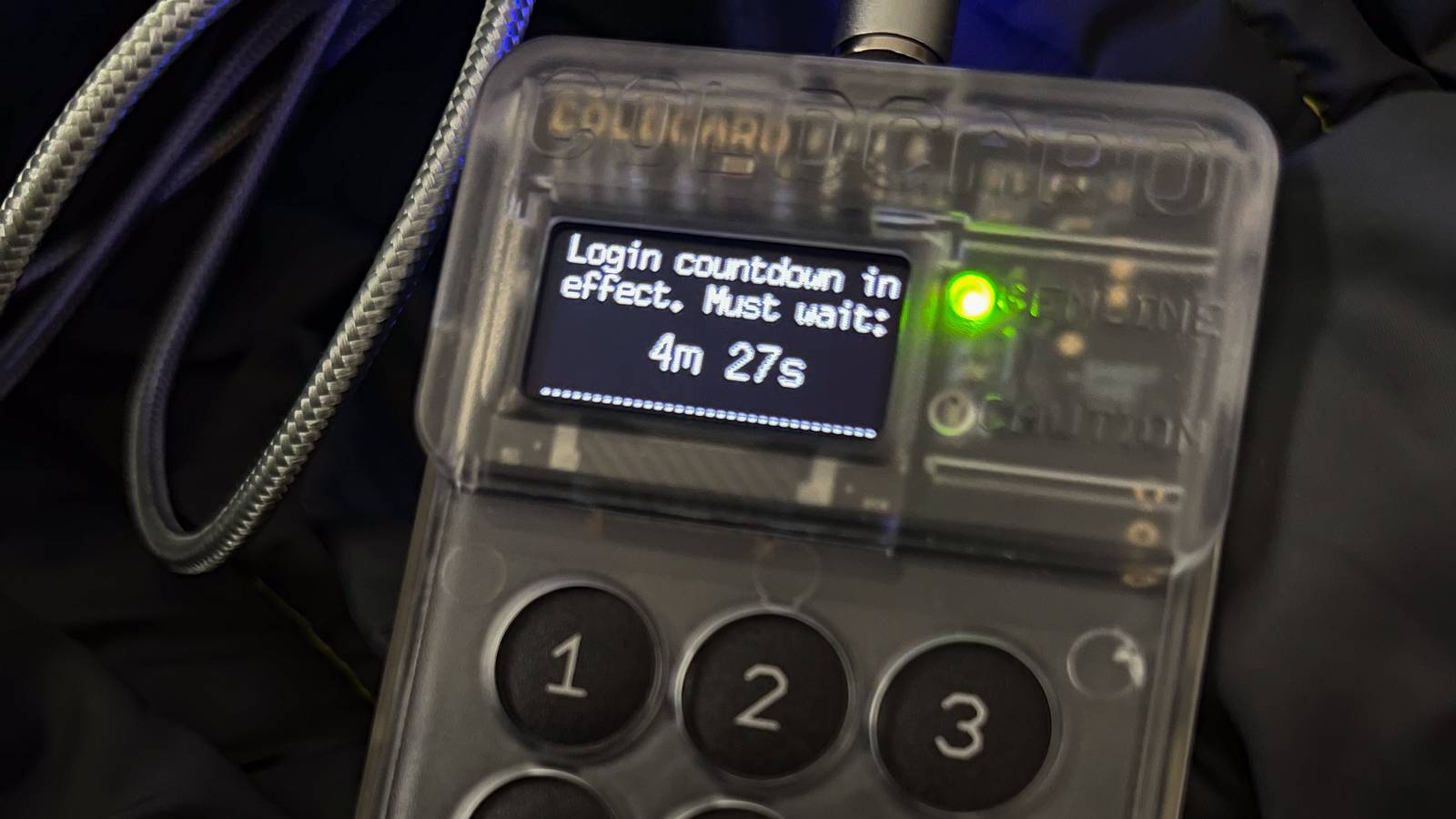 A cryptocurrency hardware wallet with a small screen and a numbered keypad, plugged into a cable