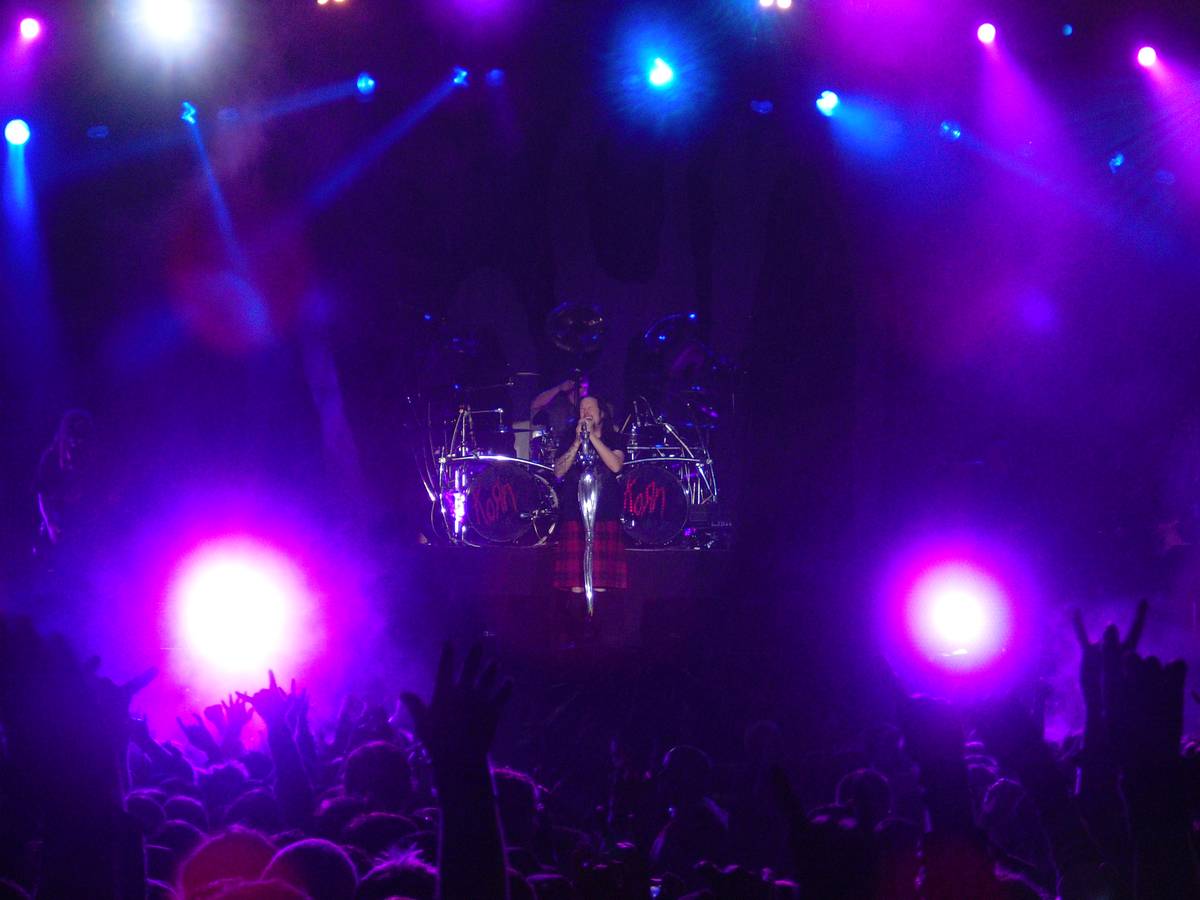 Korn's singer in a kilt at a mic stand in front of the drum kit, in purple light above a crowd with raised hands