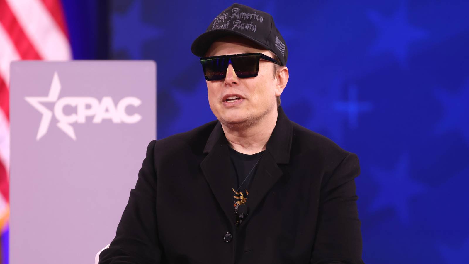 Elon Musk in a black cap, sunglasses and jacket seated on stage at a conference