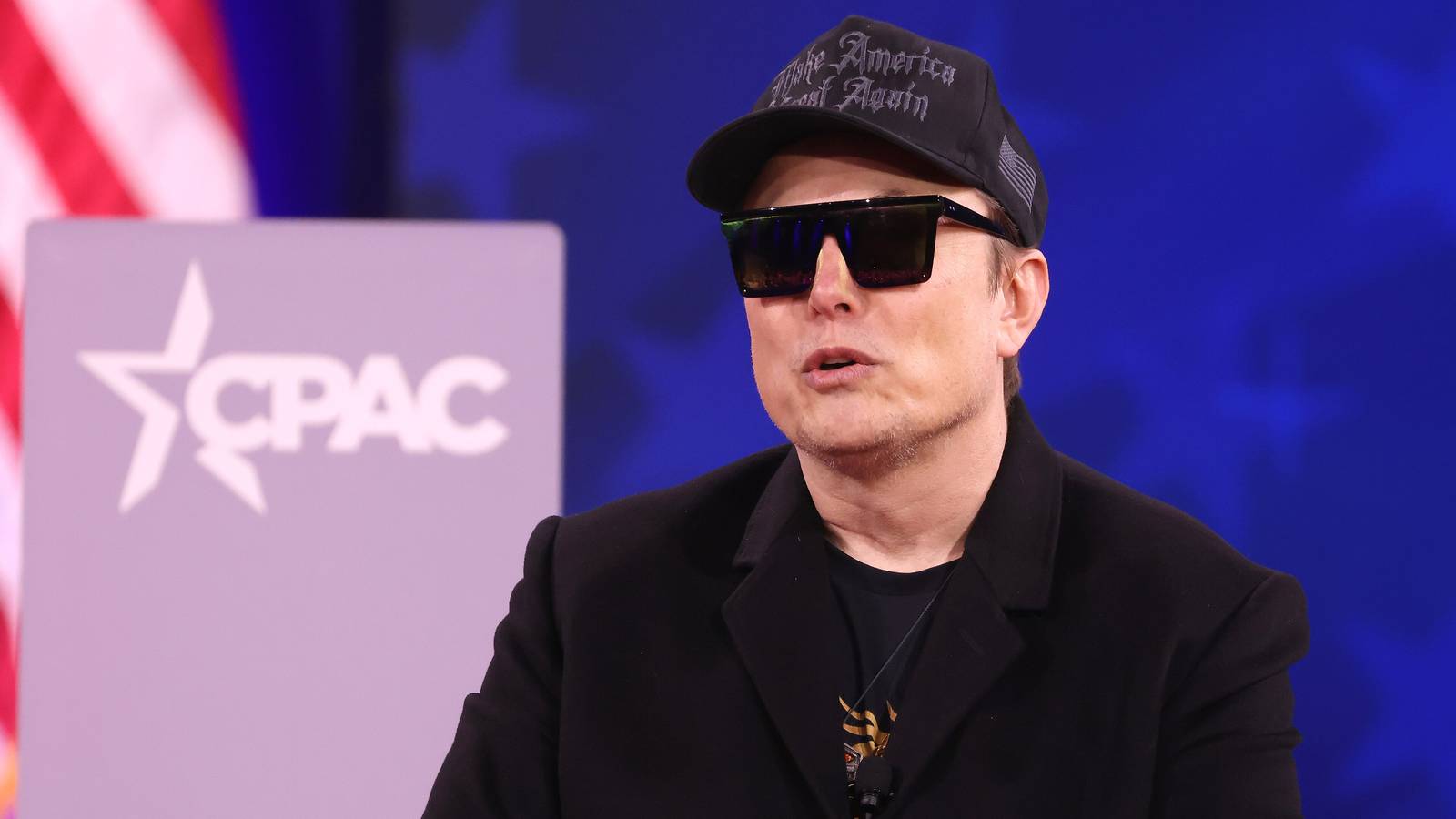 Elon Musk in a black cap, sunglasses and coat speaking on stage at CPAC