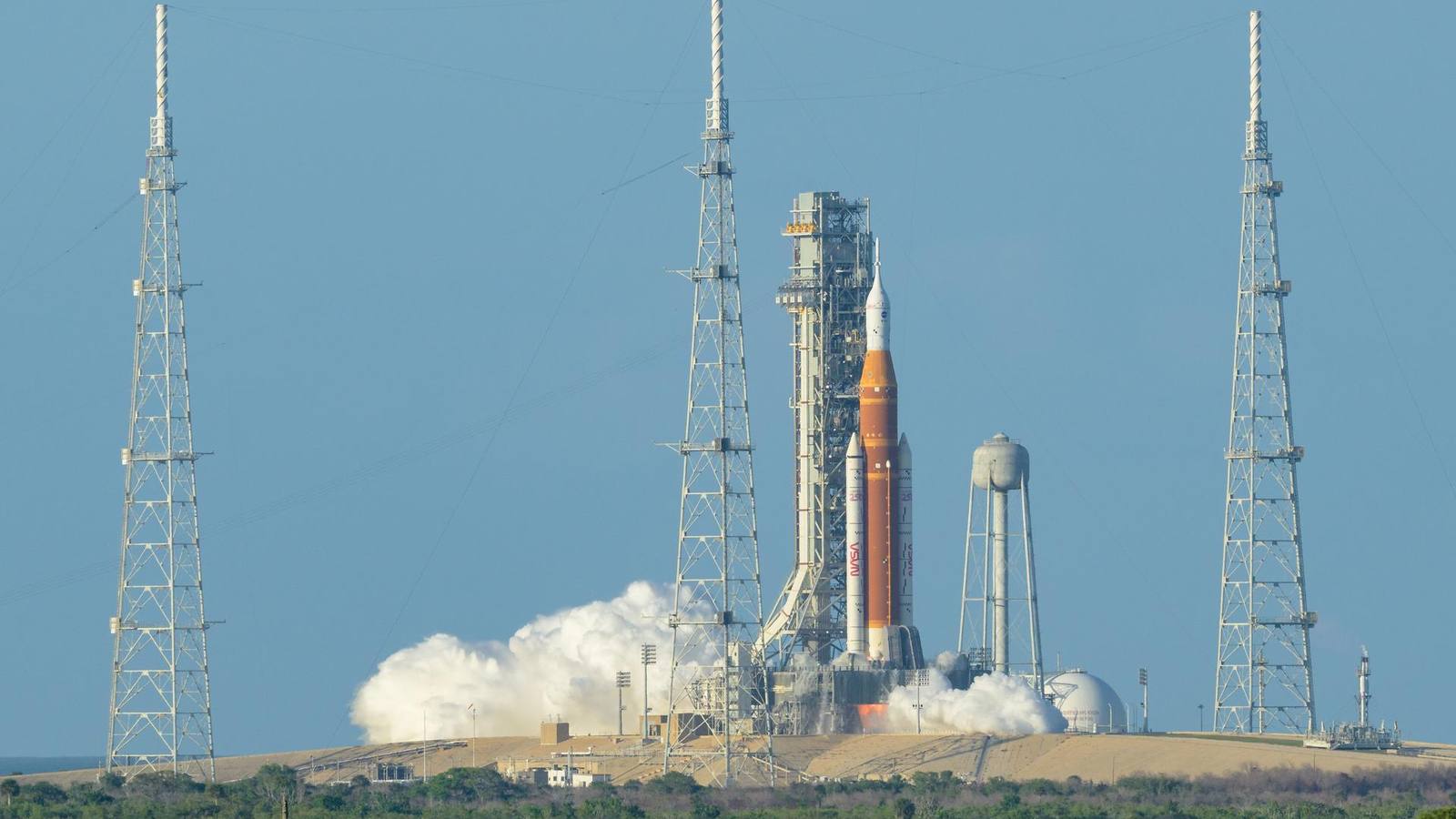 NASA's orange and white SLS rocket on the launch pad between lightning towers as clouds of steam billow at ignition
