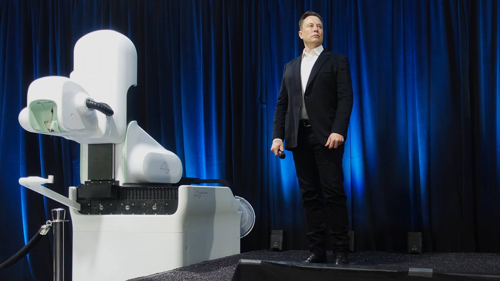 Elon Musk on stage beside Neuralink's white surgical robot in front of blue curtains at a 2020 event