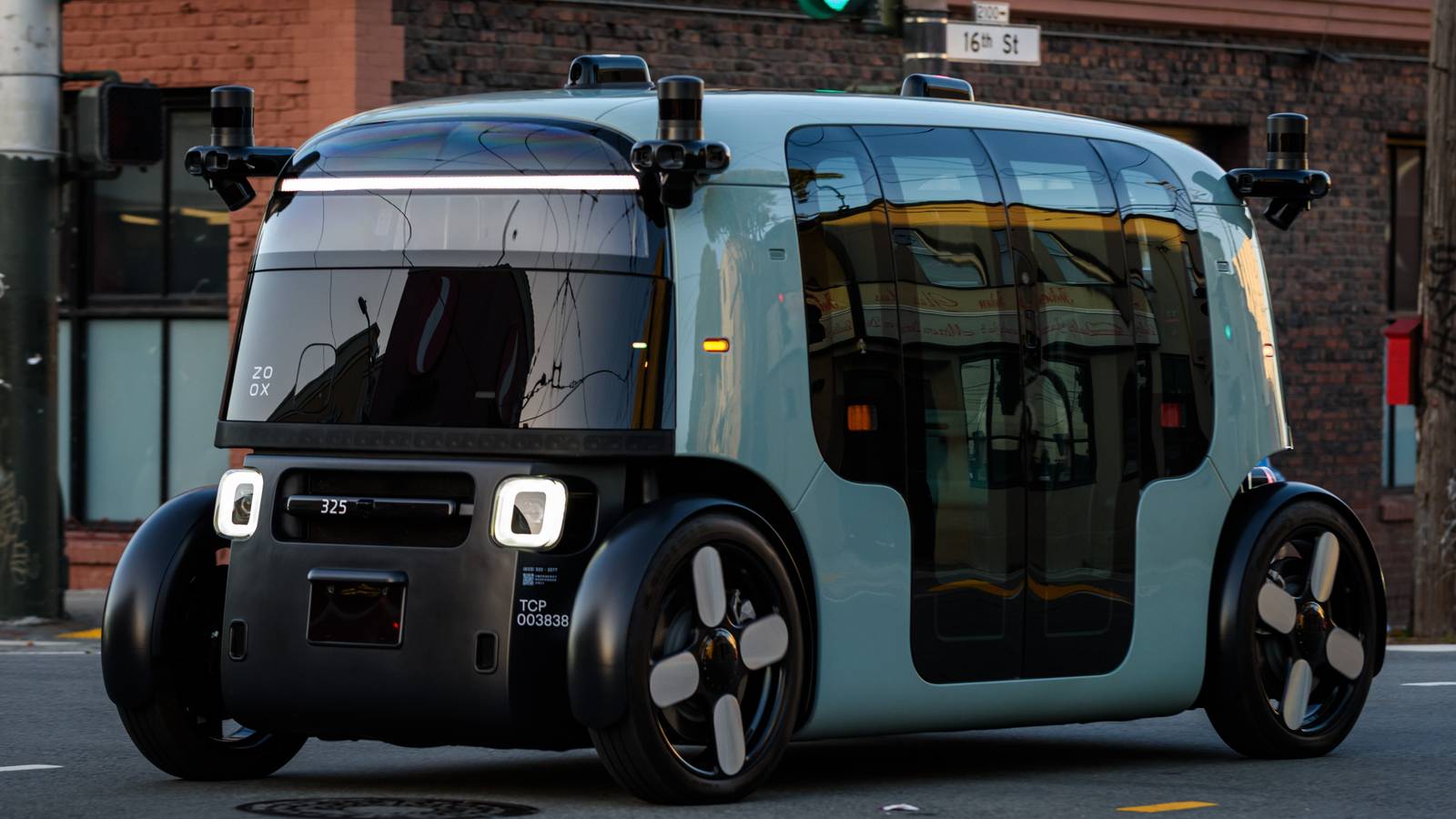 A boxy Zoox driverless robotaxi with sensor pods on its roof corners crossing a street in San Francisco