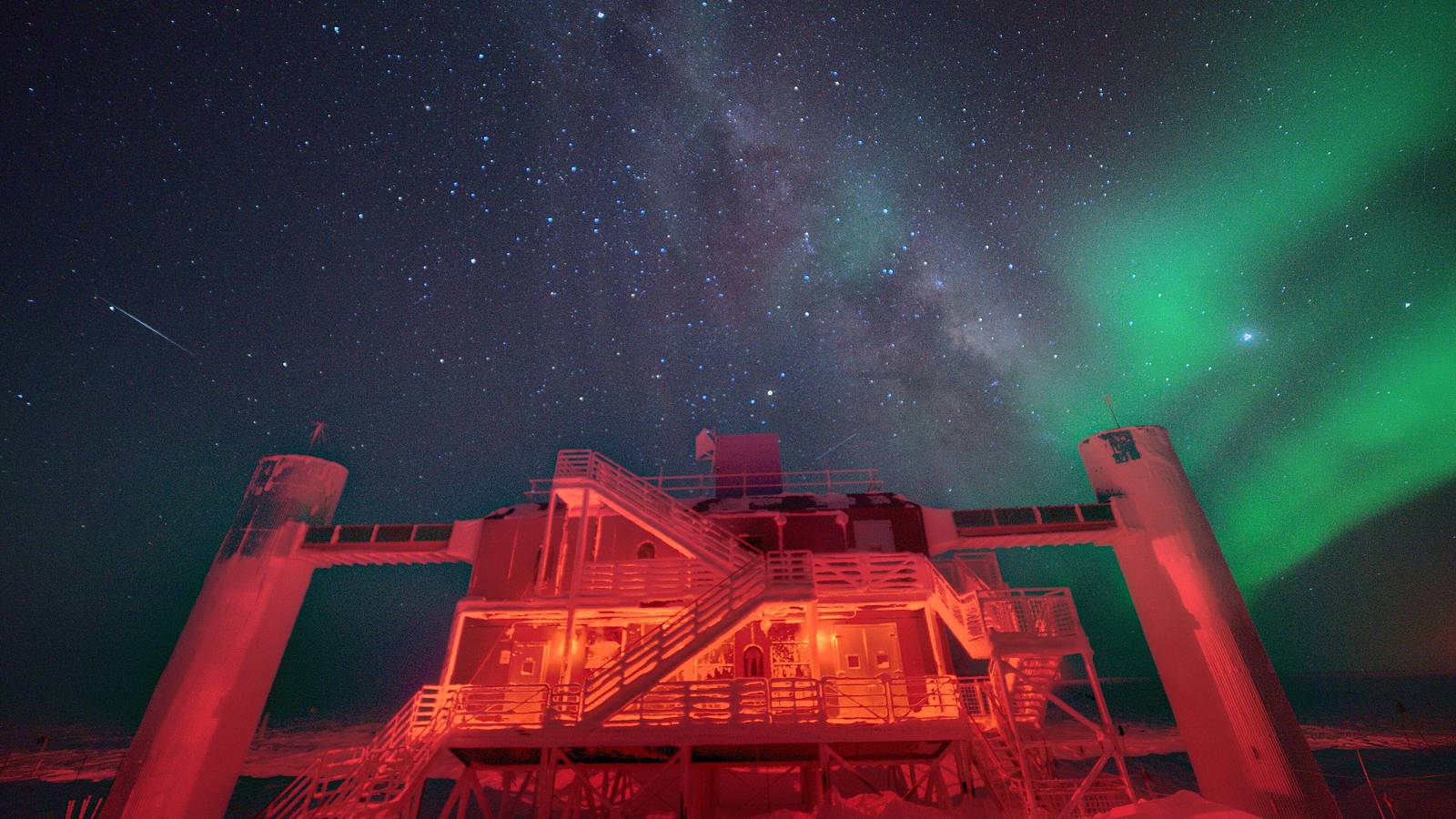 The IceCube Laboratory at the South Pole lit red under the Milky Way and a green aurora in the winter night sky
