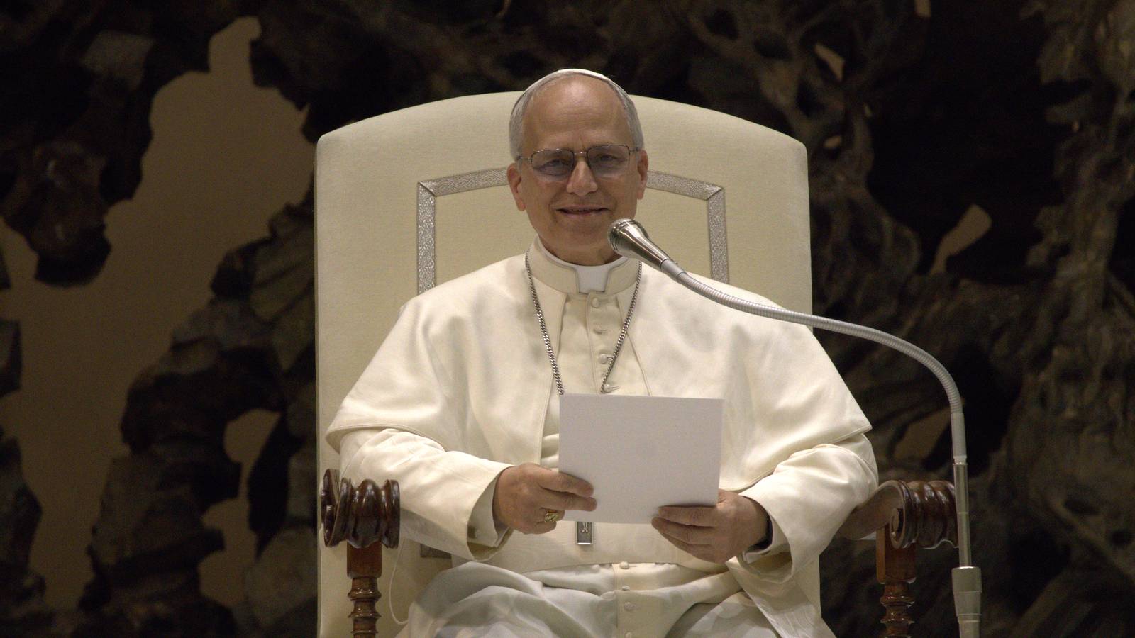 Pope Leo XIV in white papal dress smiles as he reads from a sheet of paper at a microphone