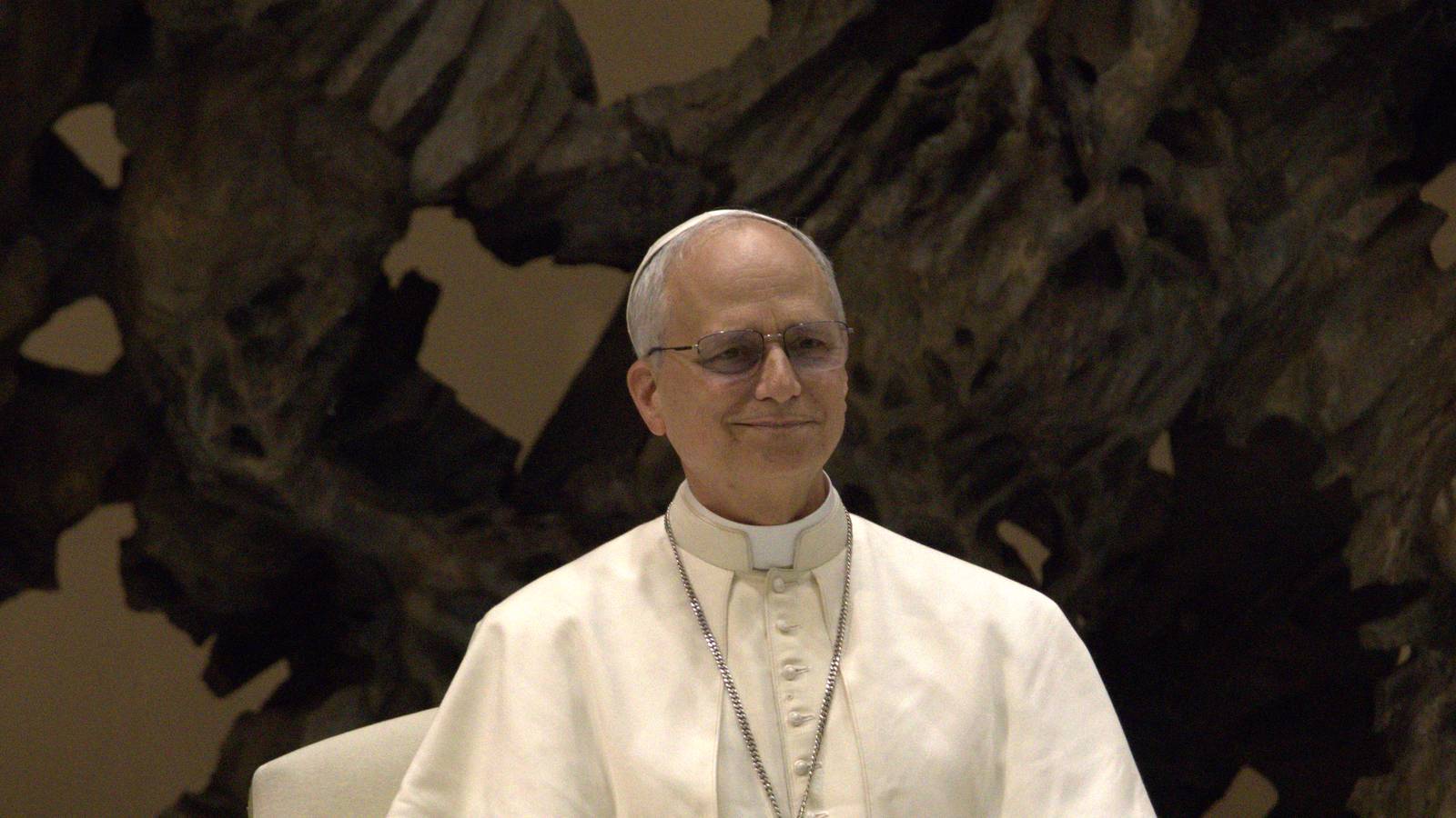 Pope Leo XIV in white robes and glasses, smiling, in front of a bronze sculpture in the Paul VI Audience Hall