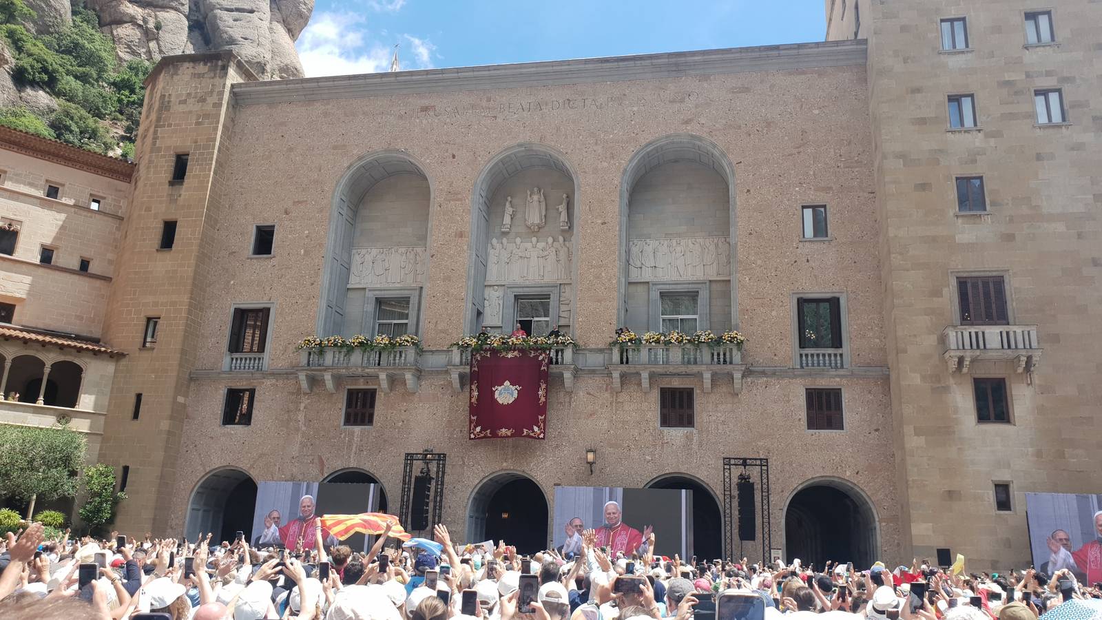 Pope Leo XIV on the abbey balcony at Montserrat as a crowd of pilgrims below watches him on big screens