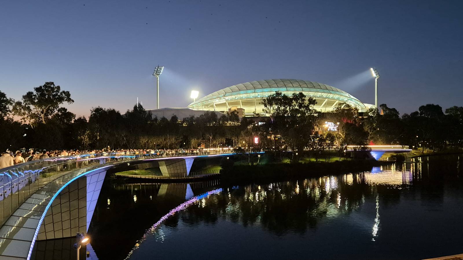 Adelaide Oval lit up at dusk beyond the Torrens footbridge and the River Torrens