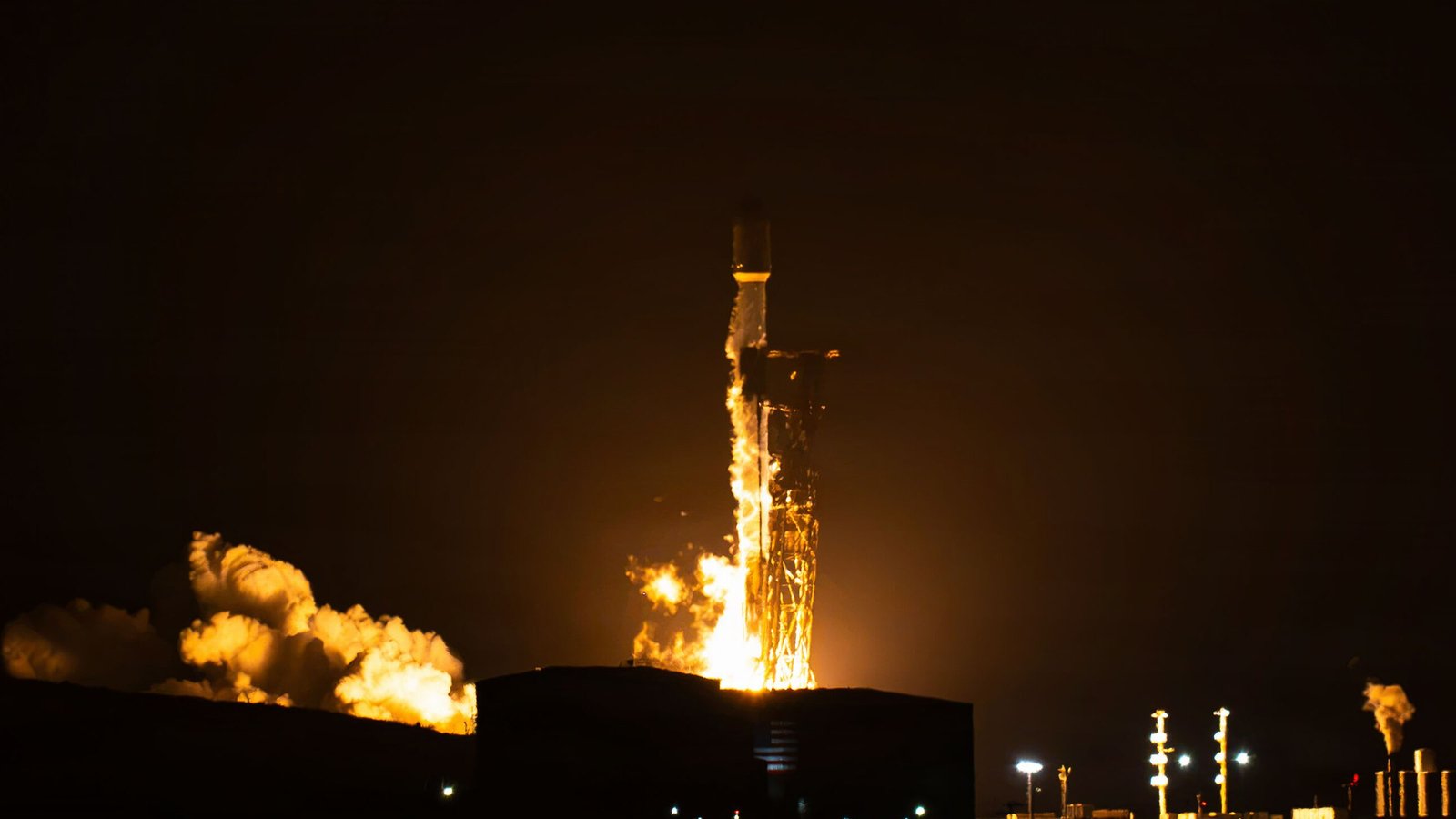 A Falcon 9 rocket lifts off at night from a Vandenberg launch pad, lighting up clouds of exhaust in orange