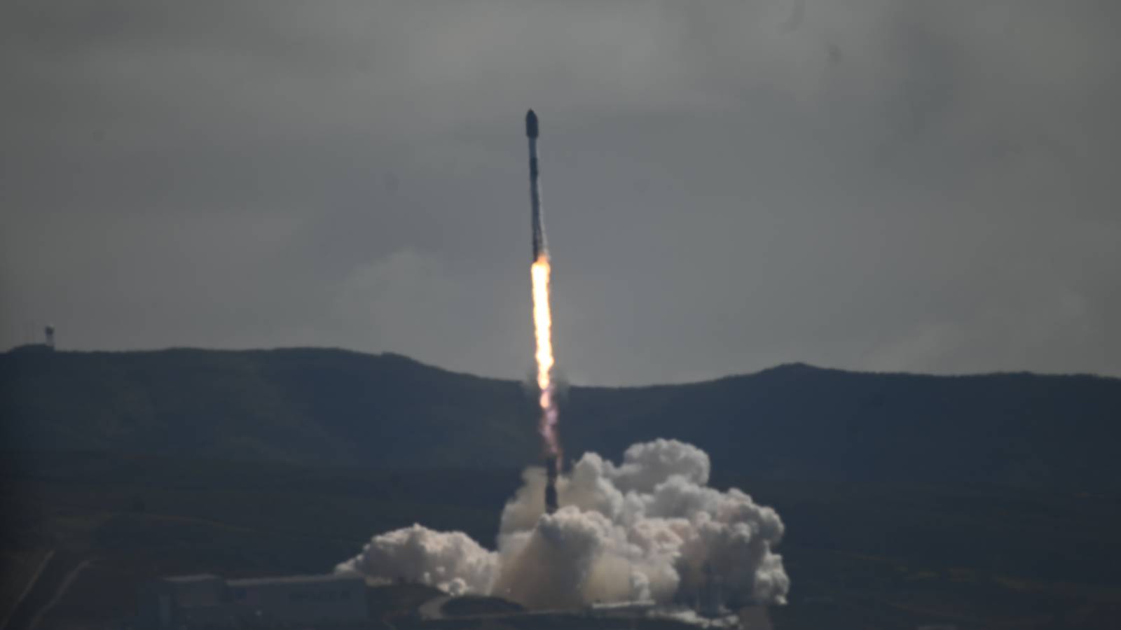 A SpaceX Falcon 9 rocket rises on a bright flame above a cloud of exhaust at Vandenberg, with dark hills under a grey sky