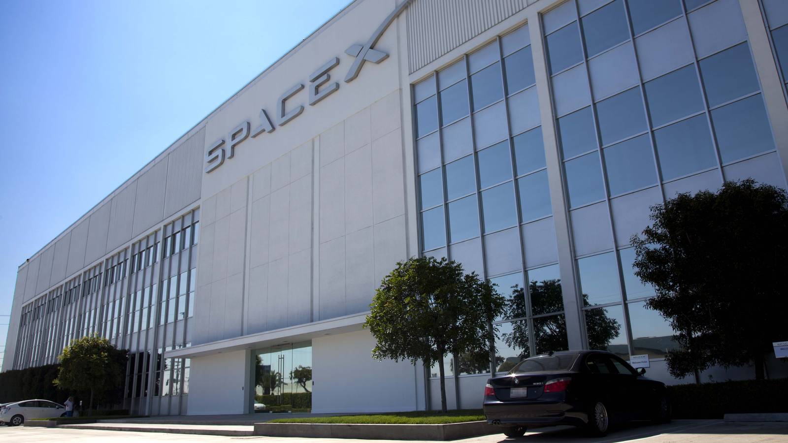The SpaceX logo on the white wall of the company's glass-fronted building in Hawthorne, California, under a blue sky