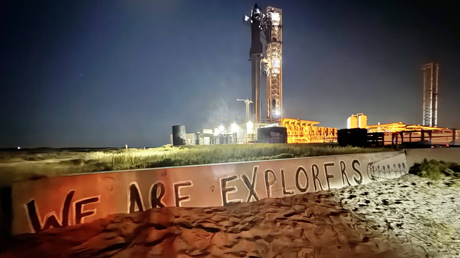 A SpaceX Starship stack lit on the Starbase pad at night behind a wall that reads We Are Explorers