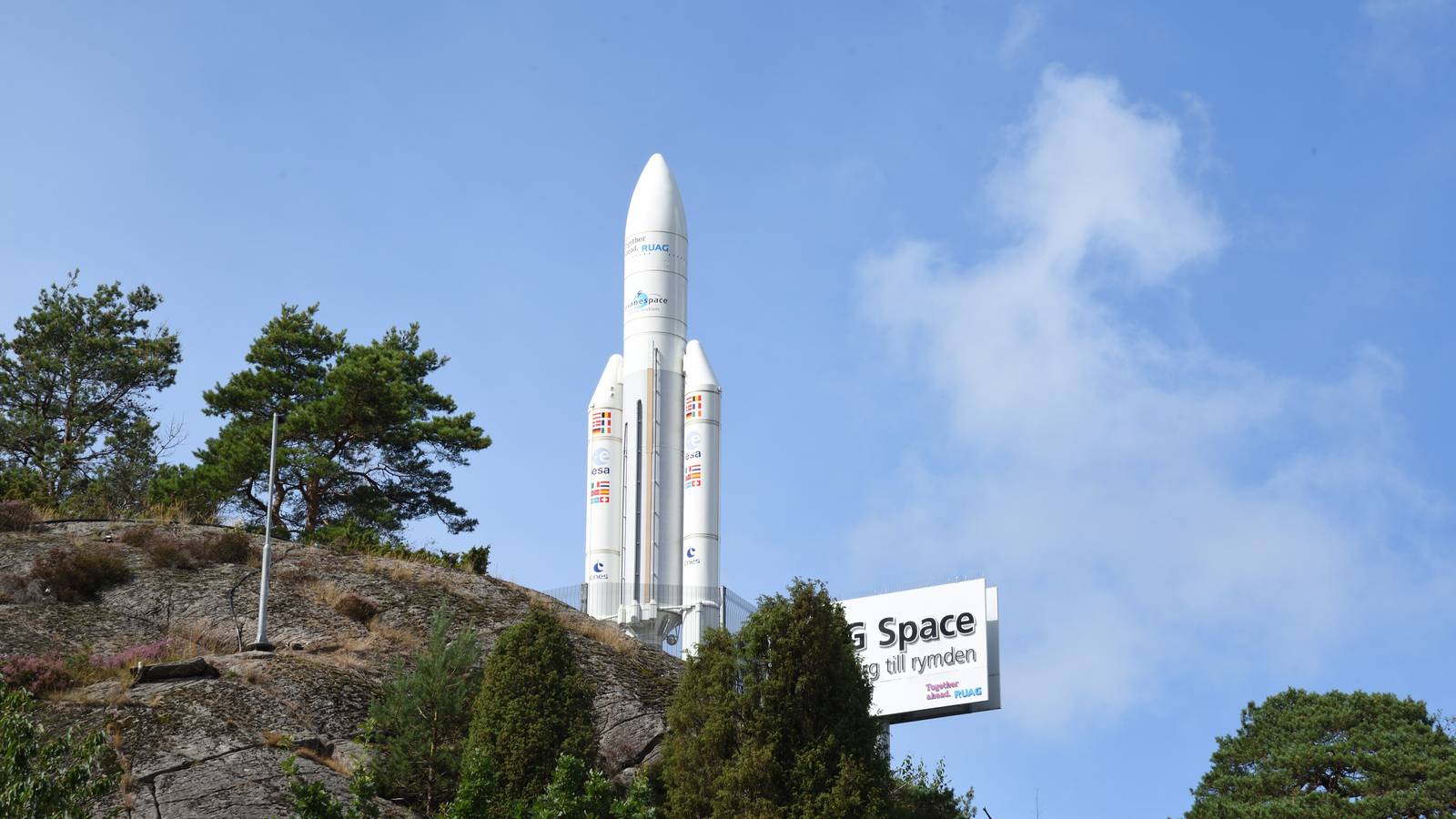 A white Ariane rocket model stands on a rocky hill beside a RUAG Space sign under a blue sky