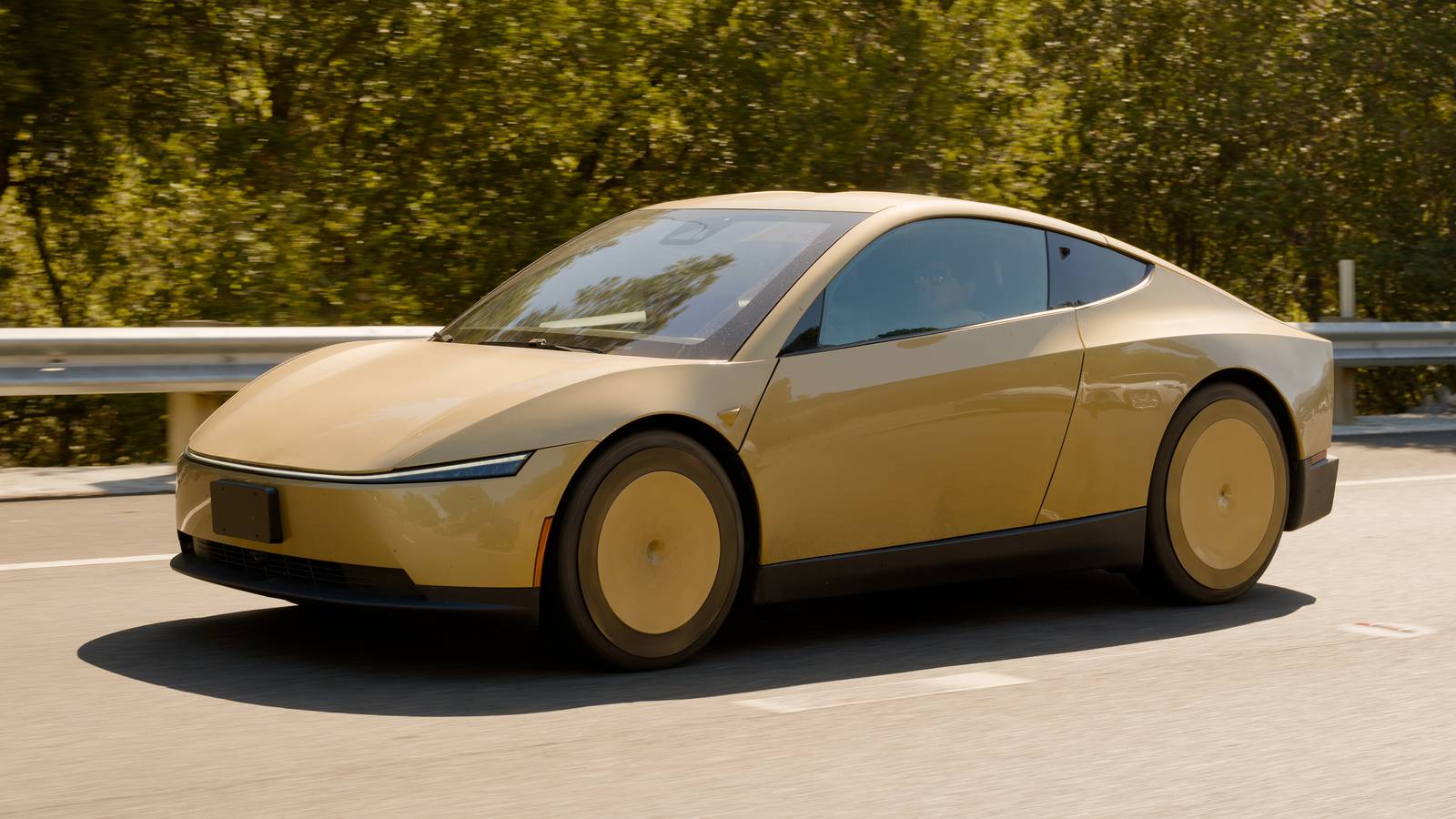 A gold Tesla Cybercab driving on a highway past a guardrail and green trees on a sunny day