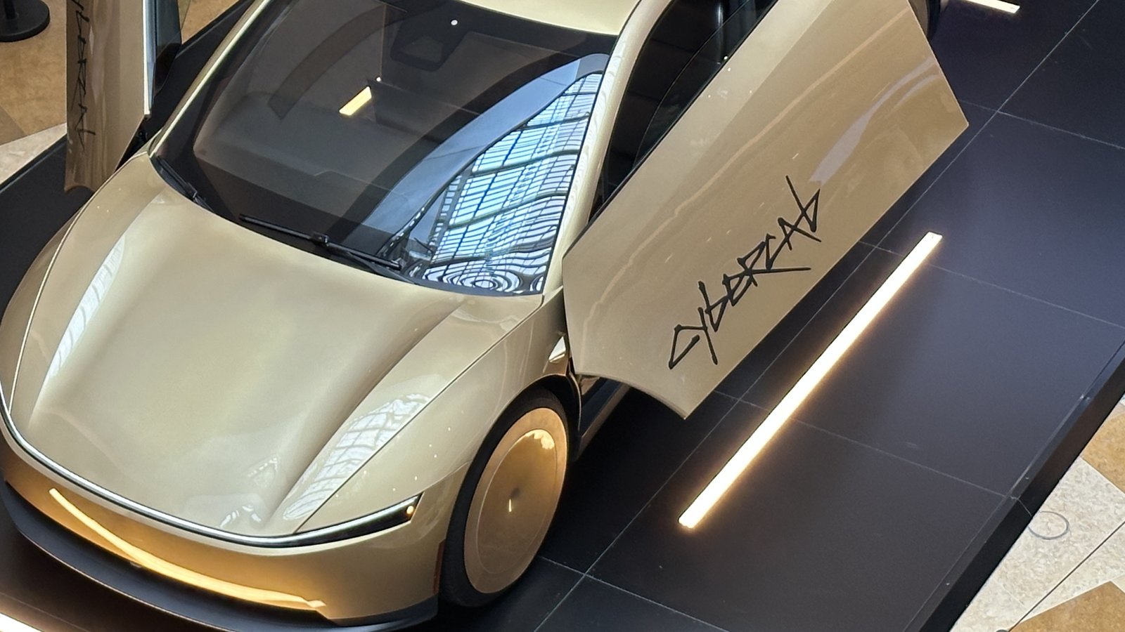 A gold Tesla Cybercab with its doors raised on a black display stand