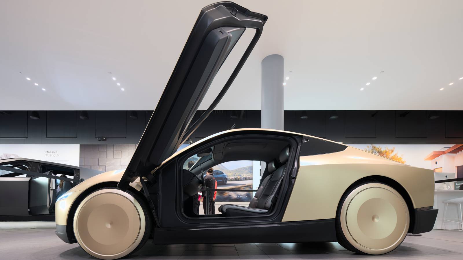 Gold Tesla Cybercab two seat robotaxi with its gull wing door raised, seen side on in a Tesla showroom