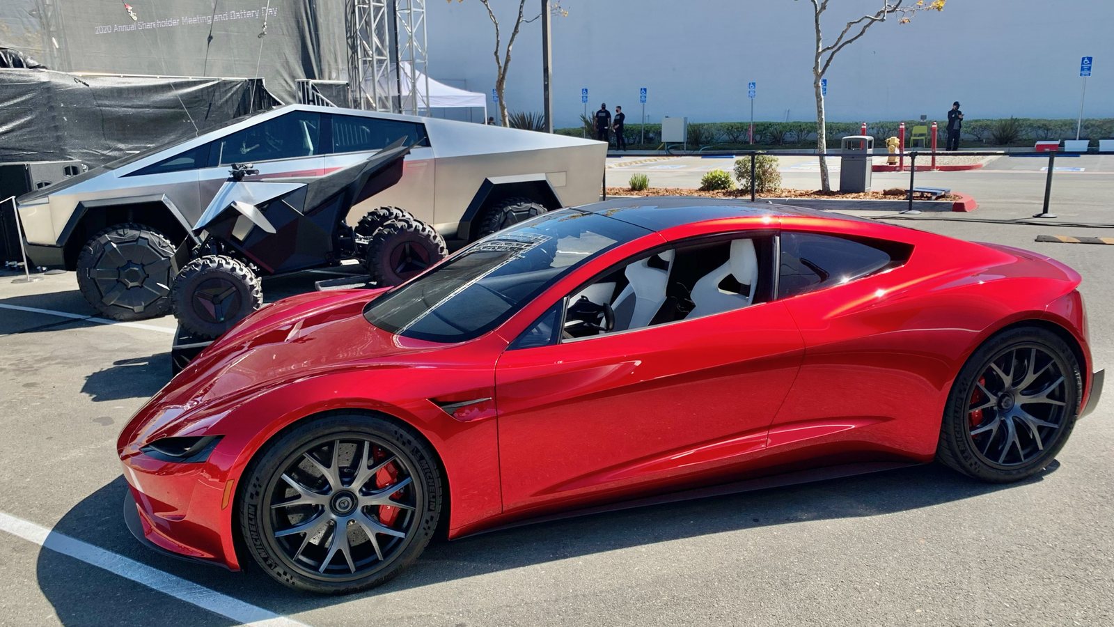 A red Tesla Roadster prototype in a sunny car park, with a Cybertruck and an electric quad bike behind it