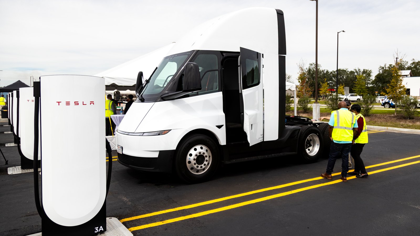 A white Tesla Semi with its door open beside a row of white Tesla Megacharger posts, with two people in hi-vis vests nearby