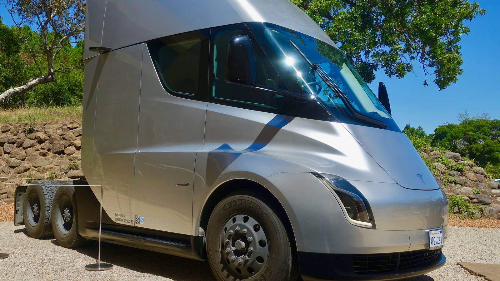 A silver Tesla Semi parked outdoors under a clear blue sky, with a stone wall and trees behind it