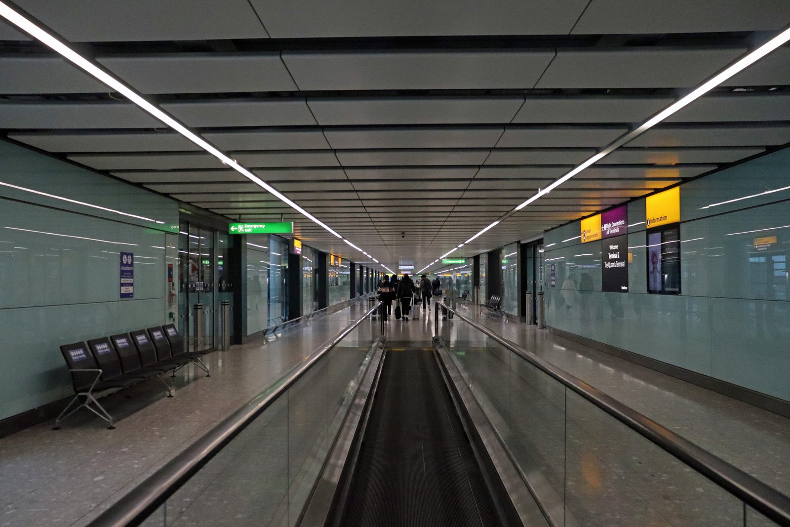 A long arrivals corridor with a moving walkway at Heathrow Terminal 2, with a few travellers walking into the distance