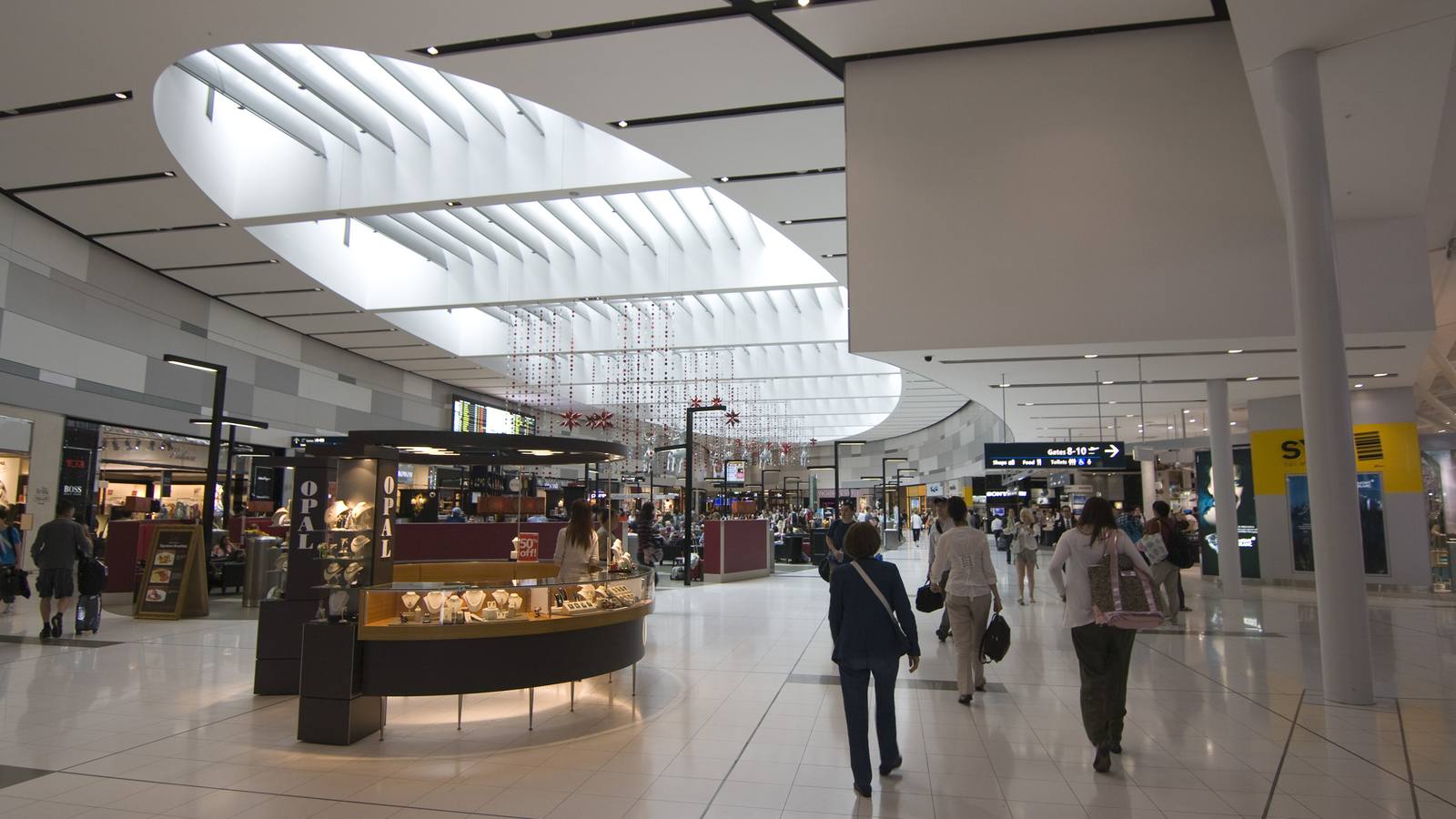 Travellers with bags walking through the bright shopping area of an airport departures hall under a curved skylight