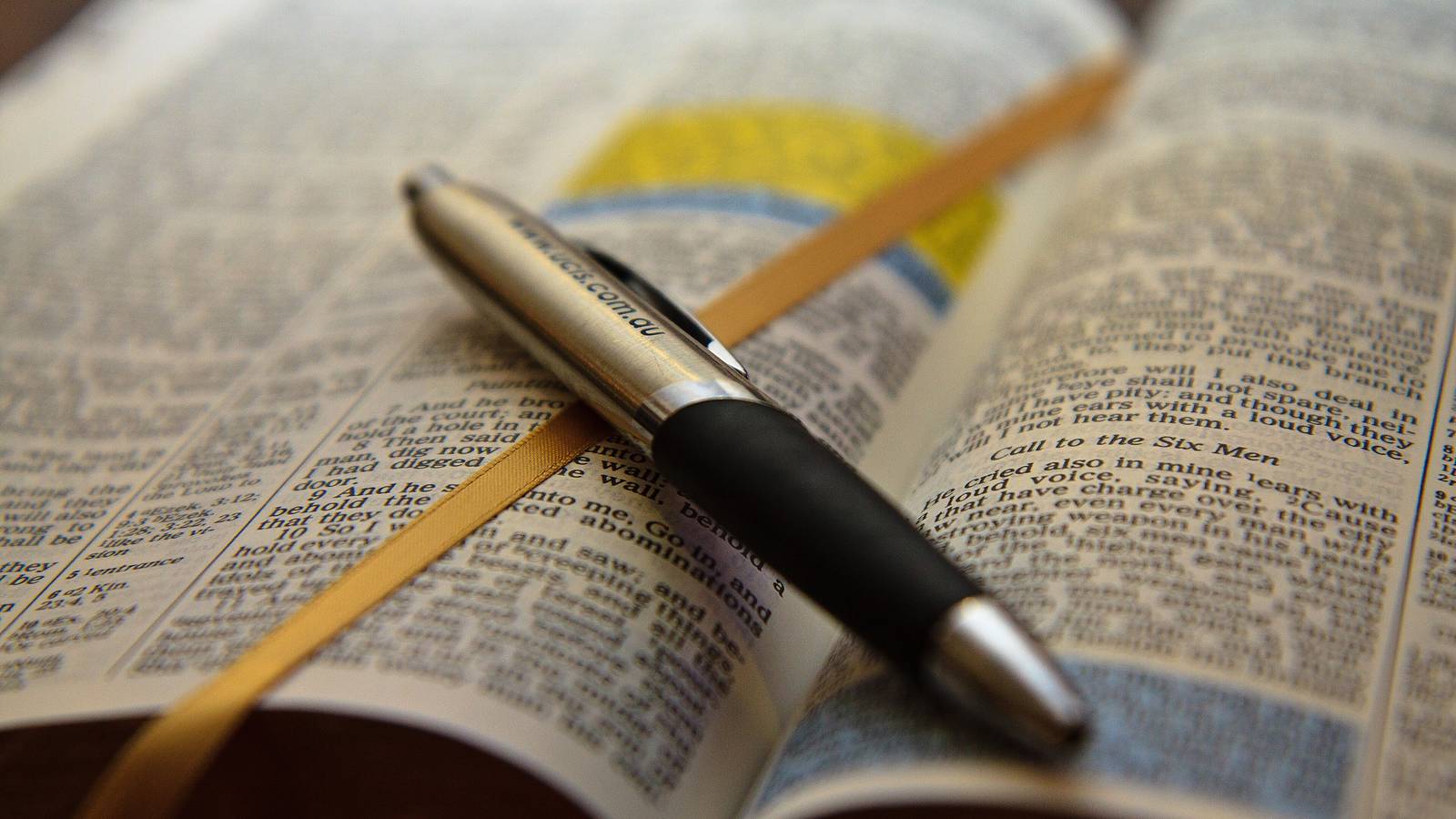 An open Bible with a highlighted passage, a gold ribbon marker and a silver and black pen resting across the pages