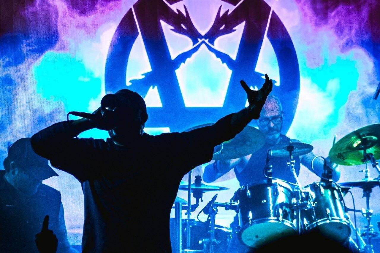 Double Dragon's singer in silhouette raises an arm onstage in front of the drummer and a glowing band logo