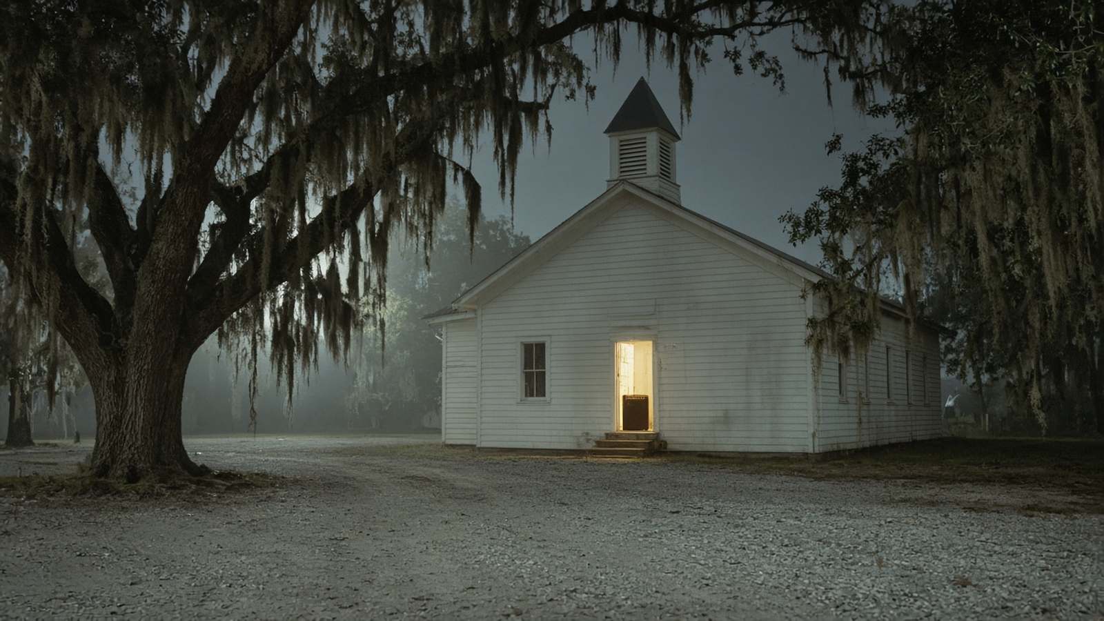 A small white clapboard church under moss-hung oaks on a misty night, its front door open and lit, with an amp just inside.